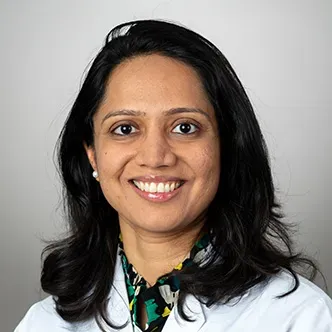 Dr. Shah has long dark hair, is wearing a white medical coat
