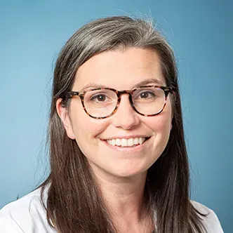 Dr. Woll has long brown hair, is wearing glasses and a white medical coat and is smiling at the camera. 