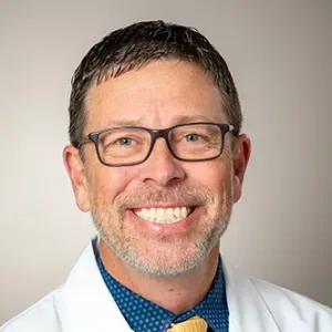 Dr. Allen has short dark hair, a graying goatee, is wearing glasses, a blue shirt and yellow tie under a white medical coat, and is smiling at the camera